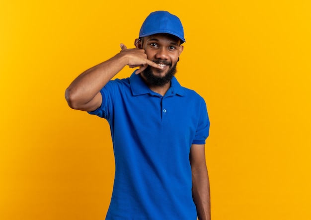 Smiling young afro-american delivery man gesturing call me sign isolated on orange background with copy space