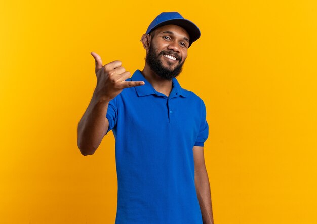 Smiling young afro-american delivery man gesturing hang loose sign isolated on orange background with copy space