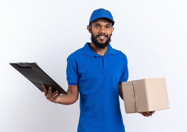 Smiling young afro-american delivery man holding cardboard box and clipboard isolated on white wall with copy space