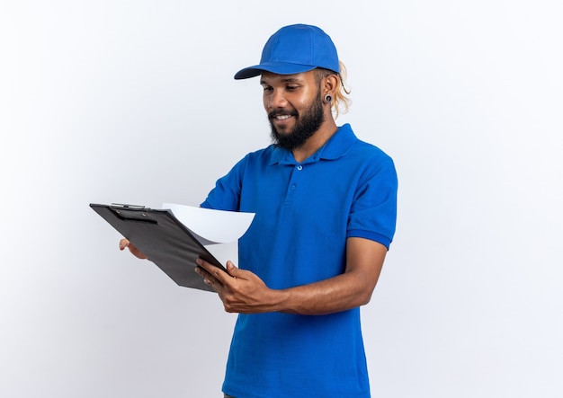 Smiling young afro-american delivery man holding and looking at clipboard isolated on white wall with copy space