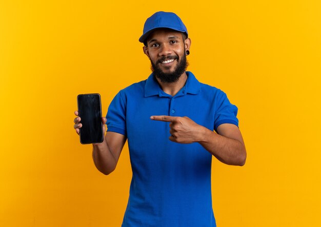 Smiling young afro-american delivery man holding and pointing at phone isolated on orange wall with copy space