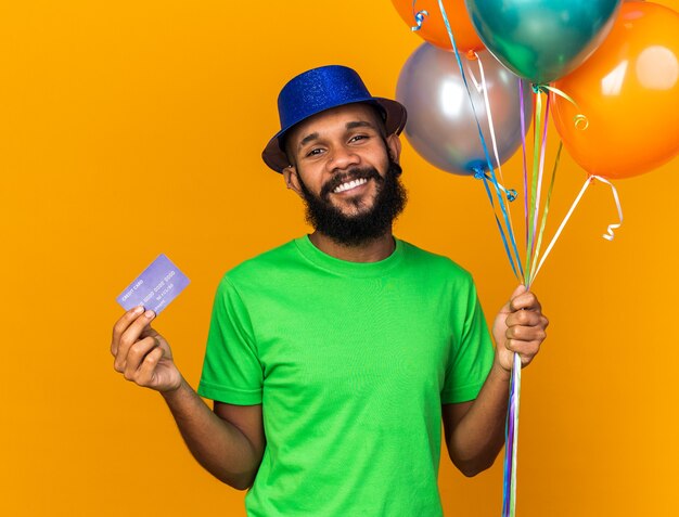 Smiling young afro-american guy wearing party hat holding balloons and credit card 