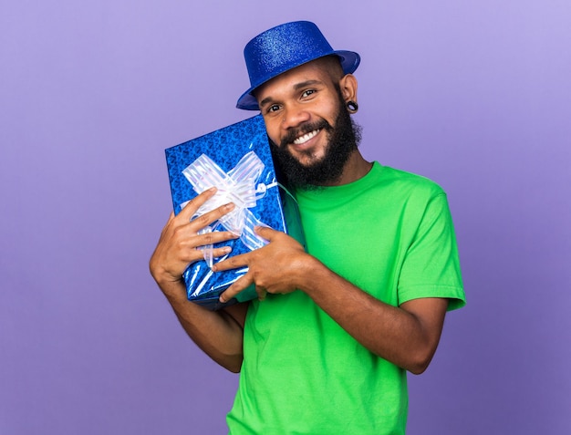 Улыбающийся молодой афро-американский парень в партийной шляпе держит подарочную коробку вокруг лица