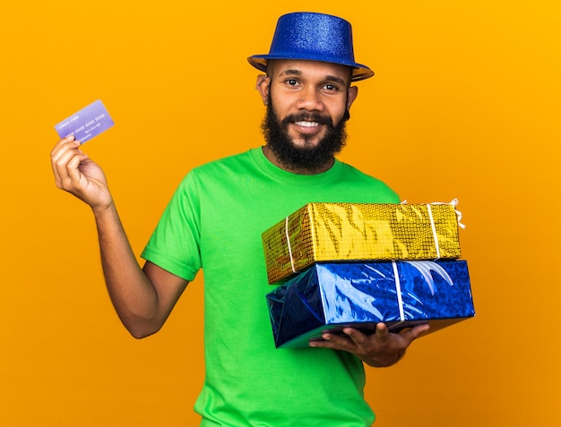 Sorridente giovane ragazzo afroamericano che indossa un cappello da festa in possesso di scatole regalo con carta di credito