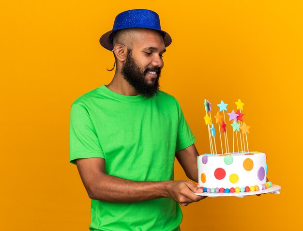 Smiling young afro-american guy wearing party hat holding and looking at cake 