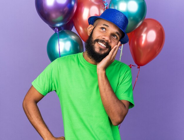 Smiling young afro-american guy wearing party hat standing in front balloons putting hand on chin 