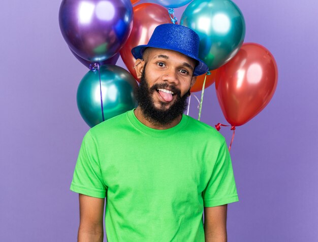 Smiling young afro-american guy wearing party hat standing in front balloons 