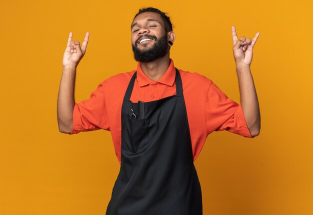 Smiling young afro-american male barber wearing uniform  doing rock sign isolated on orange wall