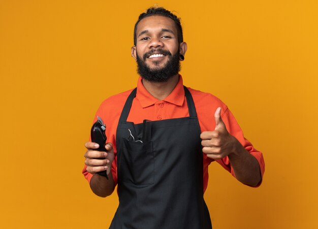 Smiling young afro-american male barber wearing uniform holding hair clippers showing thumb up 