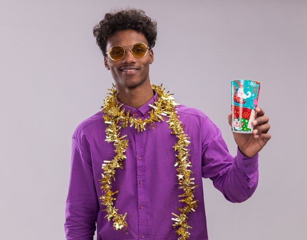 Smiling young afro-american man wearing glasses with tinsel garland around neck holding plastic christmas cup looking at camera isolated on white background