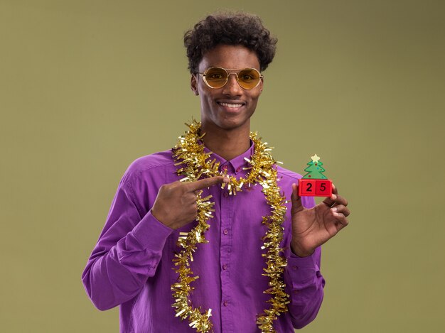 Smiling young afro-american man wearing glasses with tinsel garland around neck holding and pointing at christmas tree toy with date looking at camera isolated on olive green background