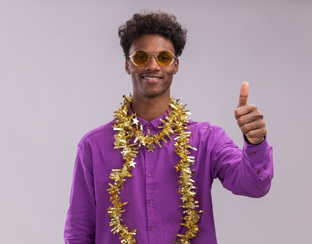 Smiling young afro-american man wearing glasses with tinsel garland around neck looking at camera showing thumb up isolated on white background