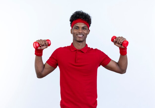 Smiling young afro-american sporty man wearing headband and wristband raising dumbbells isolated on white