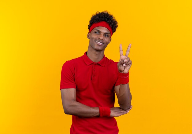 Smiling young afro-american sporty man wearing headband and wristband showing peace gesture isolated on yellow background