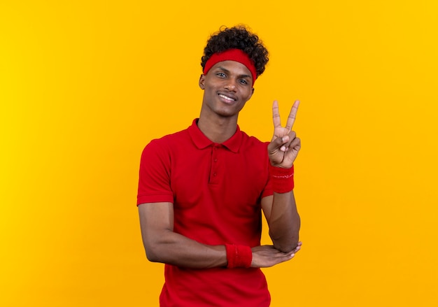 Улыбающийся молодой афро-американский спортивный мужчина с повязкой на голову и браслет, показывающий жест мира на желтом фоне