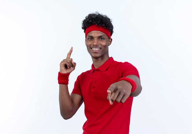 Smiling young afro-american sporty man wearing headband and wristband showing you gesture and points at up isolated on white