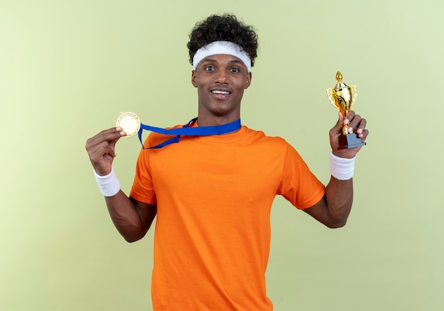 Smiling young afro-american sporty man wearing headband and wristband with medal holding cup isolated on green background