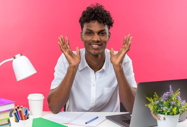 手を開いたまま学校の道具を持って机に座っている若いアフリカ系アメリカ人の学生の笑顔