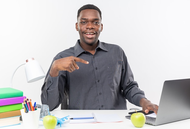 Улыбающийся молодой афро-американский студент сидит за столом со школьными принадлежностями, указывая на ноутбук
