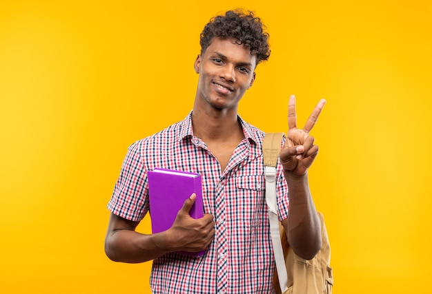 Smiling young afro-american student with backpack holding book and gesturing victory sign