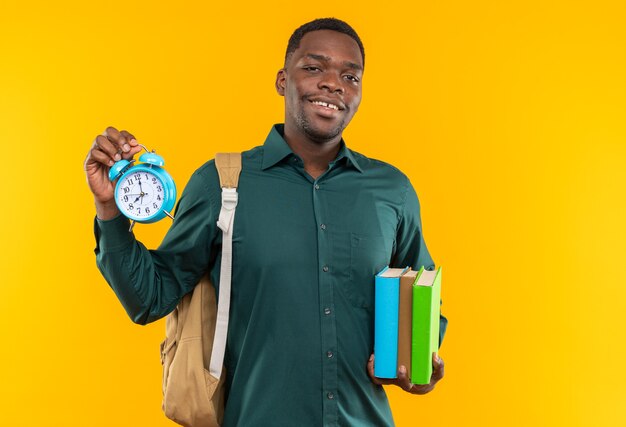 Smiling young afro-american student with backpack holding books and alarm clock