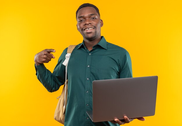 Smiling young afro-american student with backpack holding and pointing at laptop 