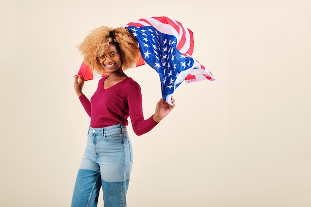 외진 배경에 서 있는 동안 미국 국기를 흔들며 웃고 있는 젊은 아프리카 여성. 미국 독립 기념일과 애국심 개념.