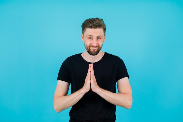 Smiling young amn is praying by holding hands together on chest on blue background