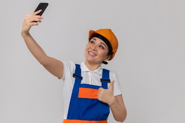 オレンジ色の安全ヘルメットをかぶって電話でselfieを取ると笑顔の若いアジアのビルダーの女の子