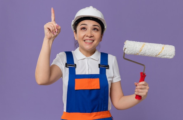 Smiling young asian builder girl with white safety helmet holding paint roller and pointing up