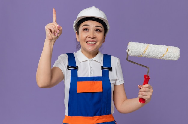 흰색 안전 헬멧을 쓰고 페인트 롤러를 들고 위를 가리키는 웃고 있는 젊은 아시아 건축업자 소녀