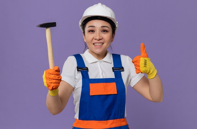 Улыбающаяся молодая азиатская женщина-строитель с белыми защитными шлемами и перчатками, держа молоток и пальцем вверх