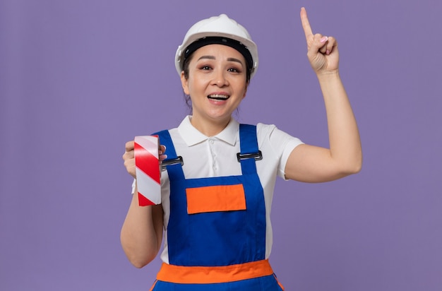 Sorridente giovane donna asiatica costruttore con casco di sicurezza bianco che tiene il nastro di avvertimento e rivolto verso l'alto