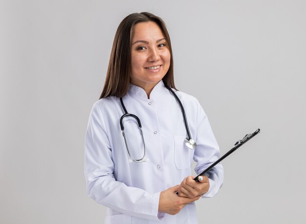 Smiling young asian female doctor wearing medical robe and stethoscope holding clipboard looking at camera isolated on white wall