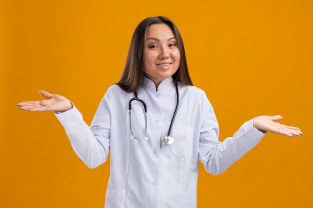 주황색 벽에 격리된 앞을 바라보는 빈 손을 보여주는 의료 가운과 청진기를 입은 웃고 있는 젊은 아시아 여성 의사