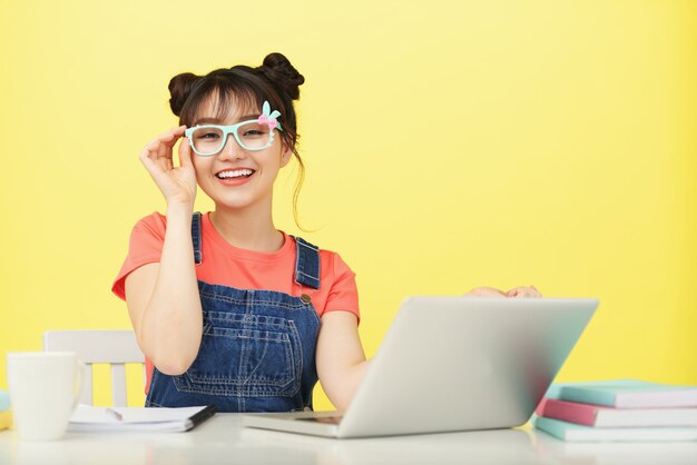 Smiling young Asian female student in brightly colored glasses sitting at desk with laptop