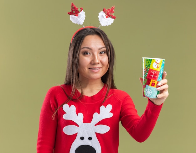 Smiling young asian girl wearing christmas hair hoop holding out christmas cup at camera isolated on olive green background