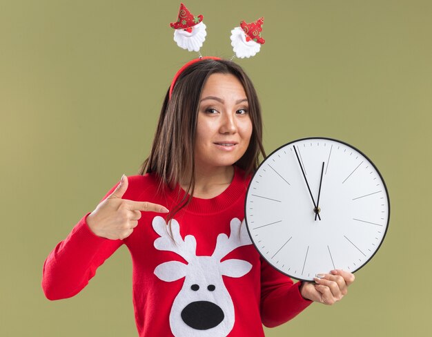 Smiling young asian girl wearing christmas hair hoop holding and points at wall clock isolated on olive green background