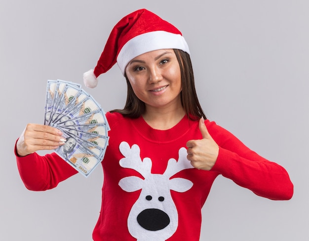 白い背景で隔離の親指を示す現金を保持しているセーターとクリスマス帽子を着て笑顔の若いアジアの女の子