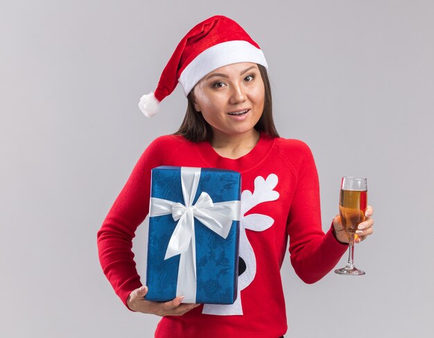 Smiling young asian girl wearing christmas hat with sweater holding gift box with glass of champagne isolated on white background