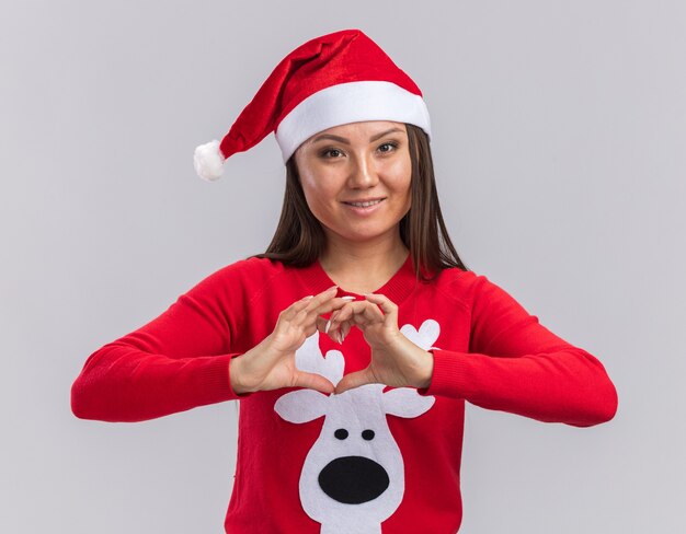Улыбающаяся молодая азиатская девушка в новогодней шапке со свитером, показывающим жест сердца на белом фоне