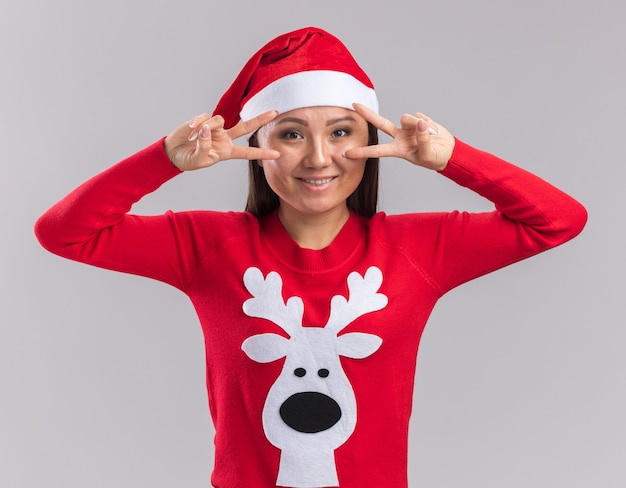 Free photo smiling young asian girl wearing christmas hat with sweater showing peace gesture isolated on white background