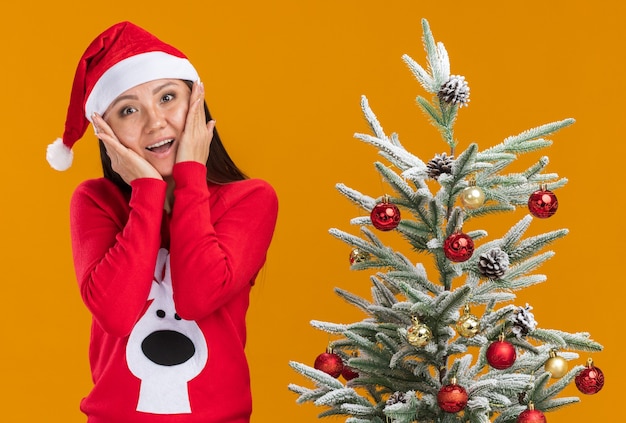 Smiling young asian girl wearing christmas hat with sweater standing nearby christmas tree putting hands on cheeks isolated on orange background