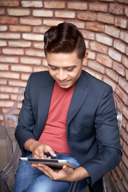 Smiling young Asian man sitting next to brick wall and using tablet