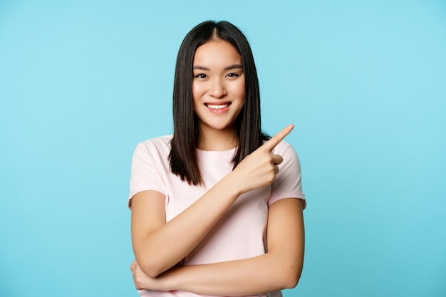 웃고 있는 20세 젊은 아시아 여성, 오른쪽 상단 모서리에 손가락을 가리키며 프로모션 배너, 파란색 배경을 보여줍니다.