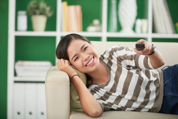 Smiling young asian woman lying on couch at home, looking straight and pressing on remote control