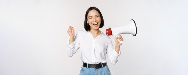 Smiling young asian woman posing with megaphone concept of news announcement and information standing over white background