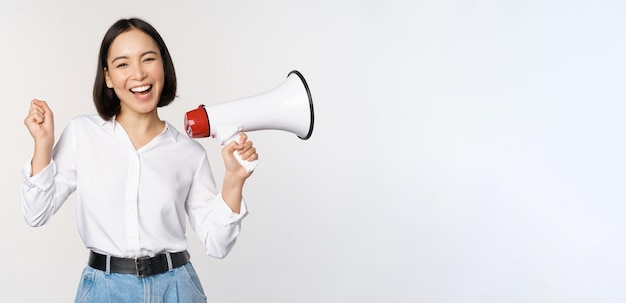 Smiling young asian woman posing with megaphone concept of news announcement and information standing over white background