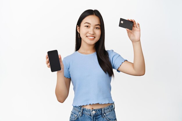 Smiling young asian woman showing credit card in raised hand empty phone screen concept of online banking and shopping white background