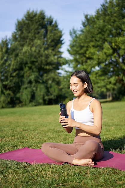 La giovane donna asiatica sorridente si siede sull'erba facendo gli esercizi nell'allenamento del parco sul prato beve l'acqua da bo